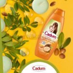 Champú Cadum de aceite de argán rodeado de hojas verdes, nueces de argán y velas en un fondo amarillo